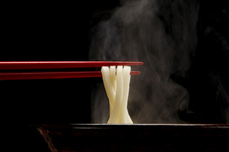 Japanese Sanuki Udon, fat straight noodles lifted out of the soup bowl with red chopsticks.  Vapor looks sharp against a black background.  Eye level perspective.