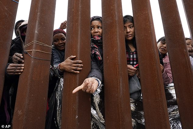 Migrants reach through a border wall for clothing distributed by volunteers while waiting between two border walls to receive an asylum application in San Diego on Friday, May 12, 2023.  The number of migrants crossing the border has fallen by about 50 percent since the end of Title 42 authority.  Biden said things were going 'better than you all expected'