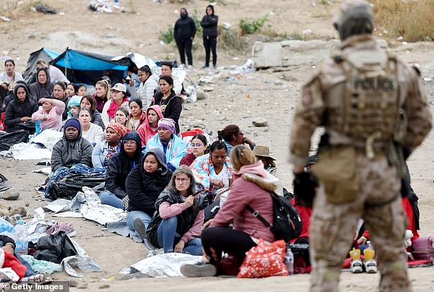 Immigrants seeking asylum in the U.S., stuck at a makeshift camp between U.S.-Mexico border walls, sit while a Customs and Border Protection officer stands guard as other migrants queue for transportation May 13, 2023